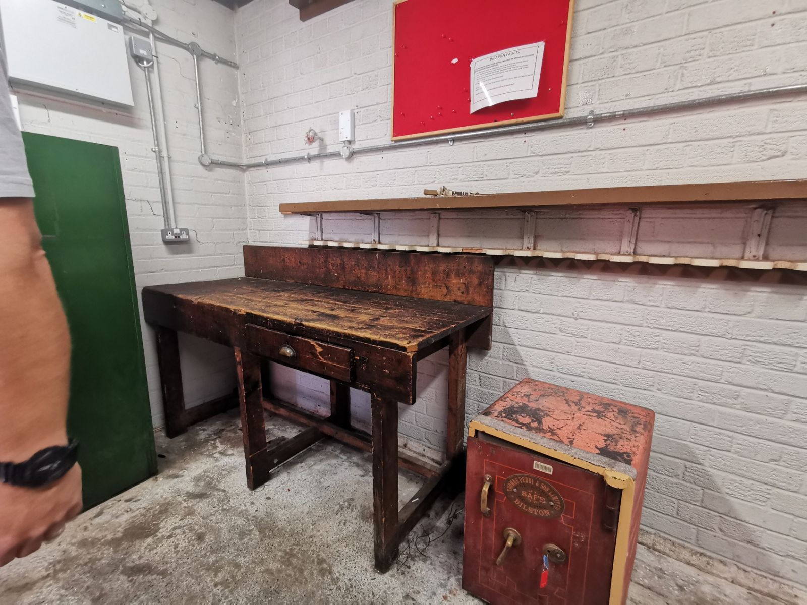 Dark wooden workbench beside an antique red safe against a white painted brick wall