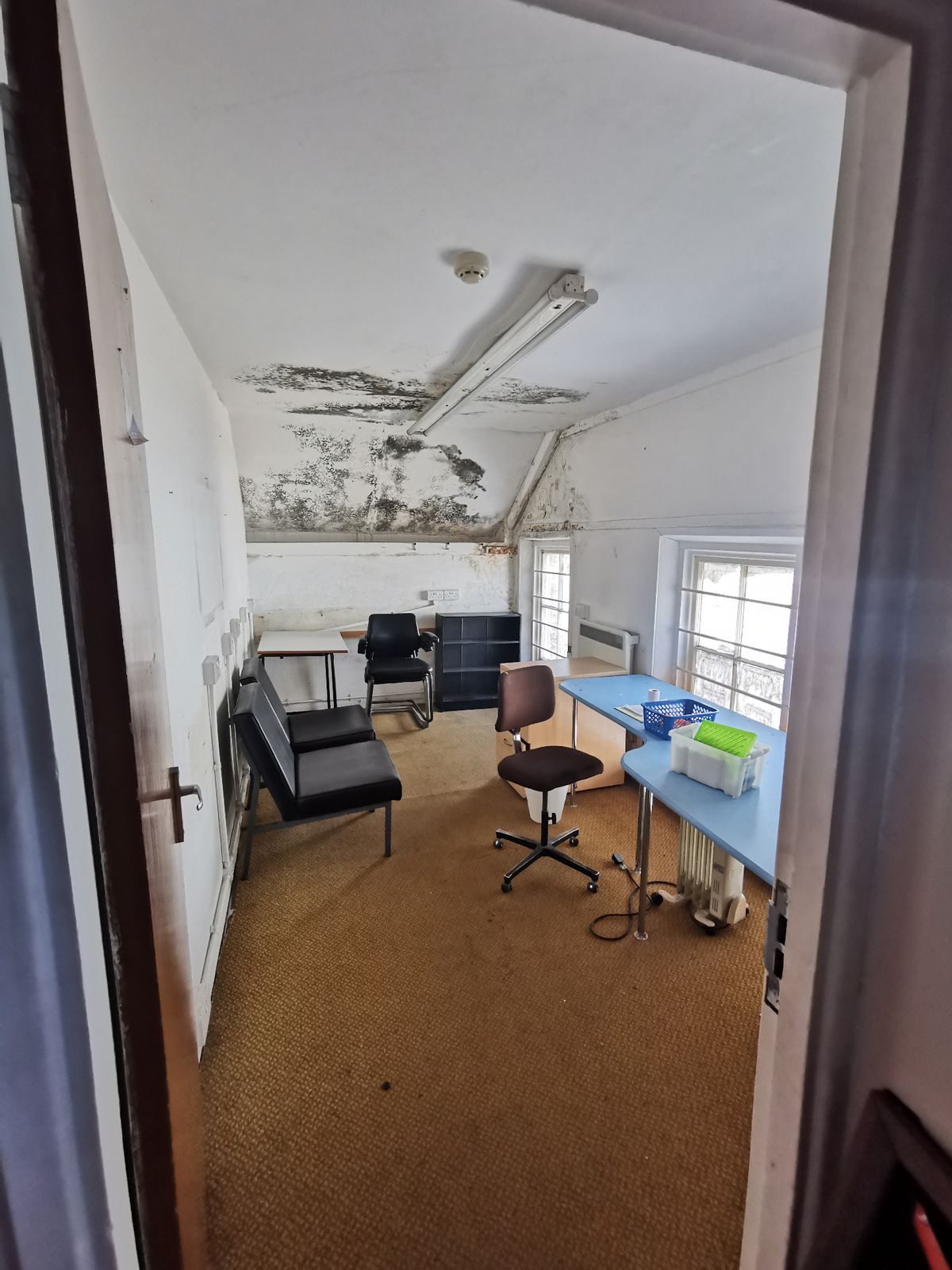 Small room with a damaged ceiling and mould, furnished with chairs, desks and a blue table